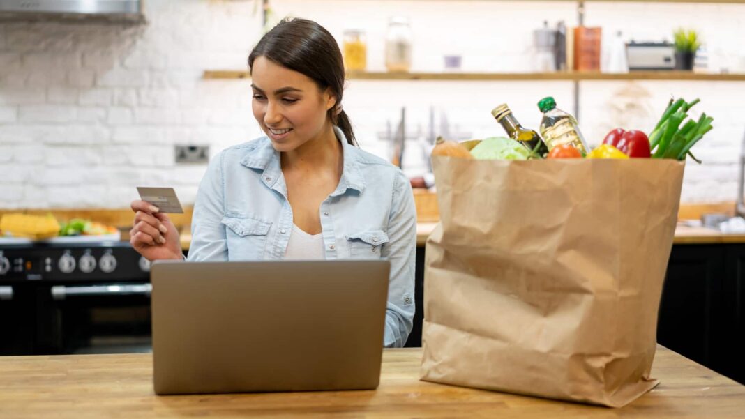 woman online shopping next to bag of groceries