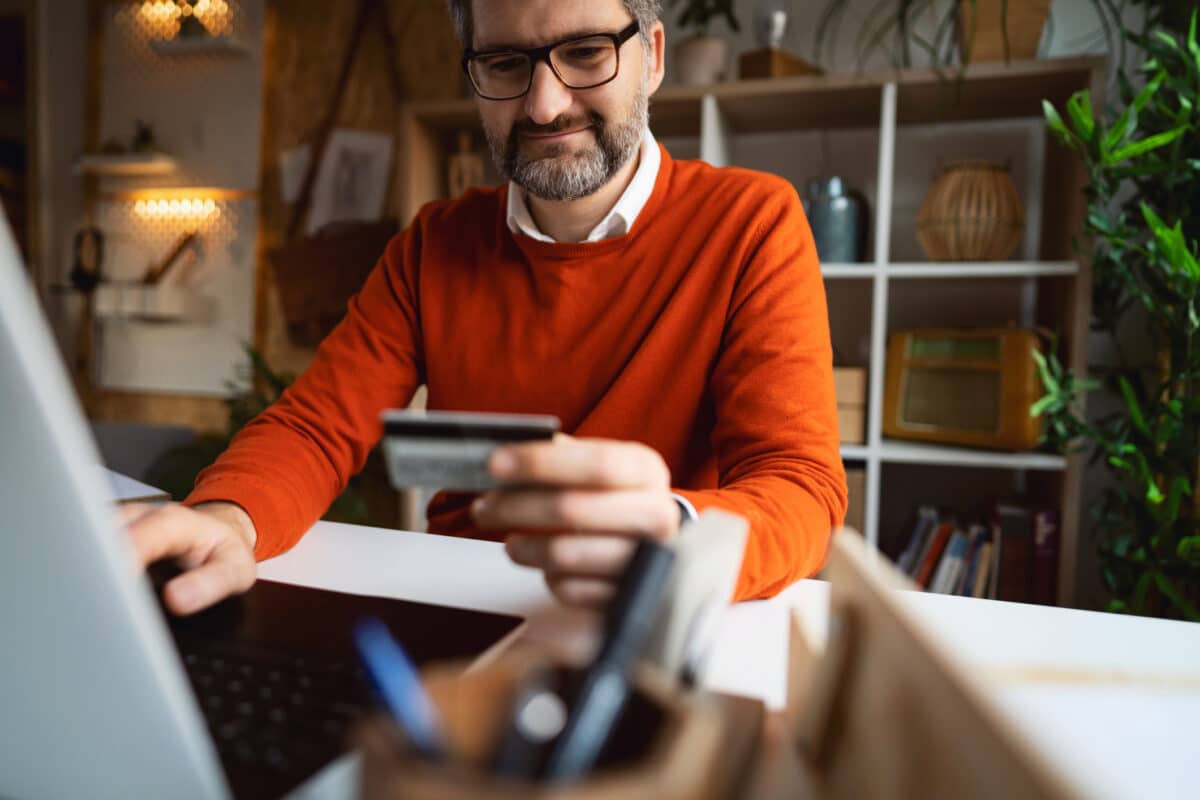 Middle aged white man wearing an orange sweater, smiling, and holding a credit card while looking at his laptop