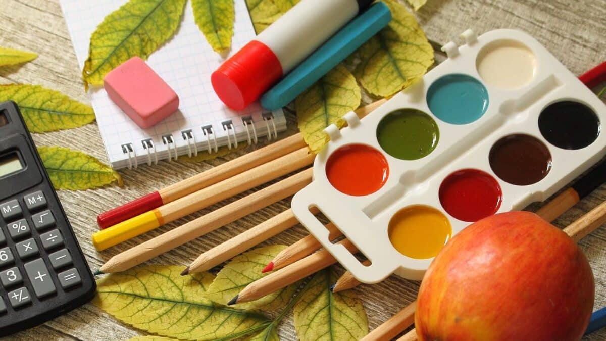 Paint, colored pencils, eraser, calculator, an apple and some green leaves scattered on a table