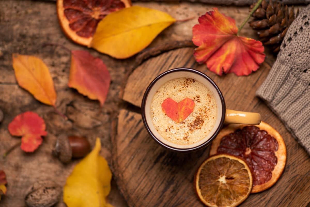 Coffee cup with orange heart on the foam, surrounded by autumn leaves, citrus slices, and a knitted sweater