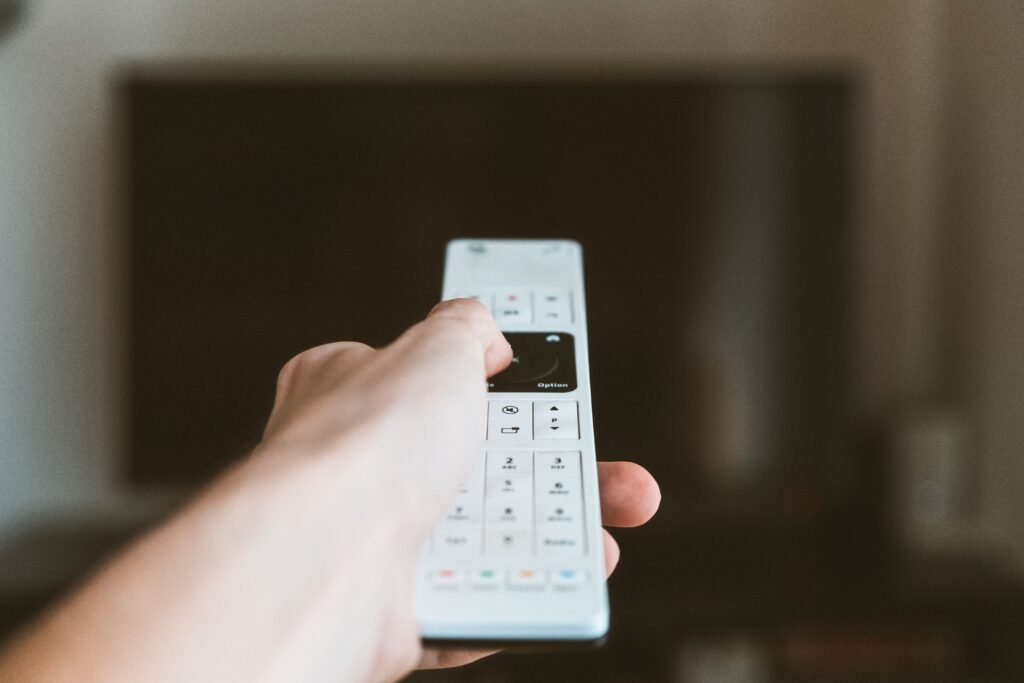 Hand holding a remote control with a TV in the background.