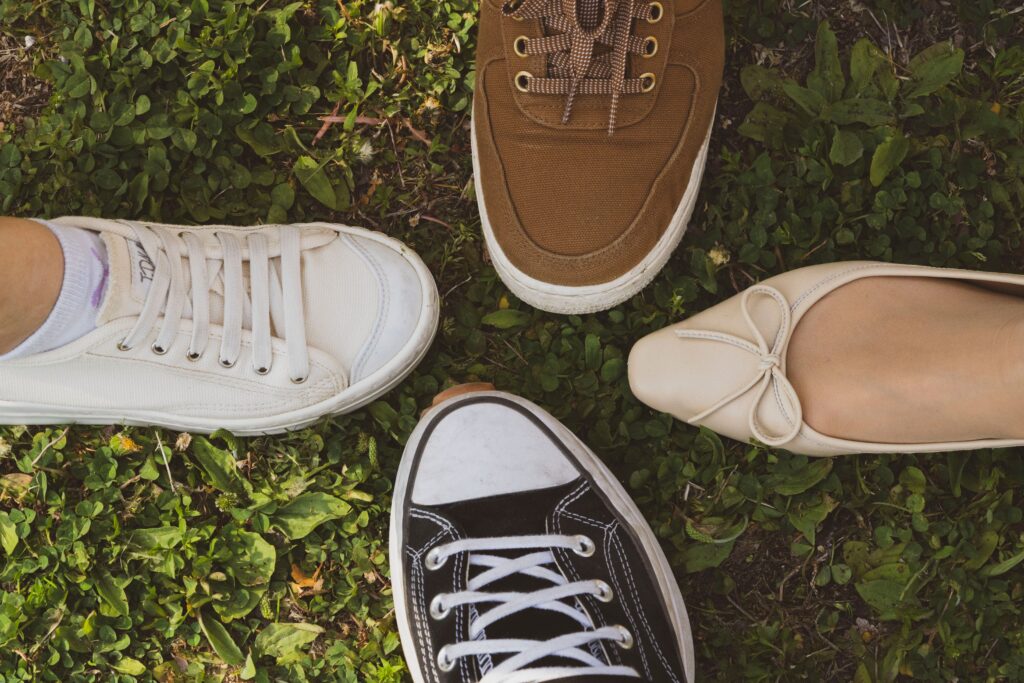 Four shoes on grass — a white sneaker, a brown canvas shoe, a black Converse-style sneaker and a beige ballet flat.