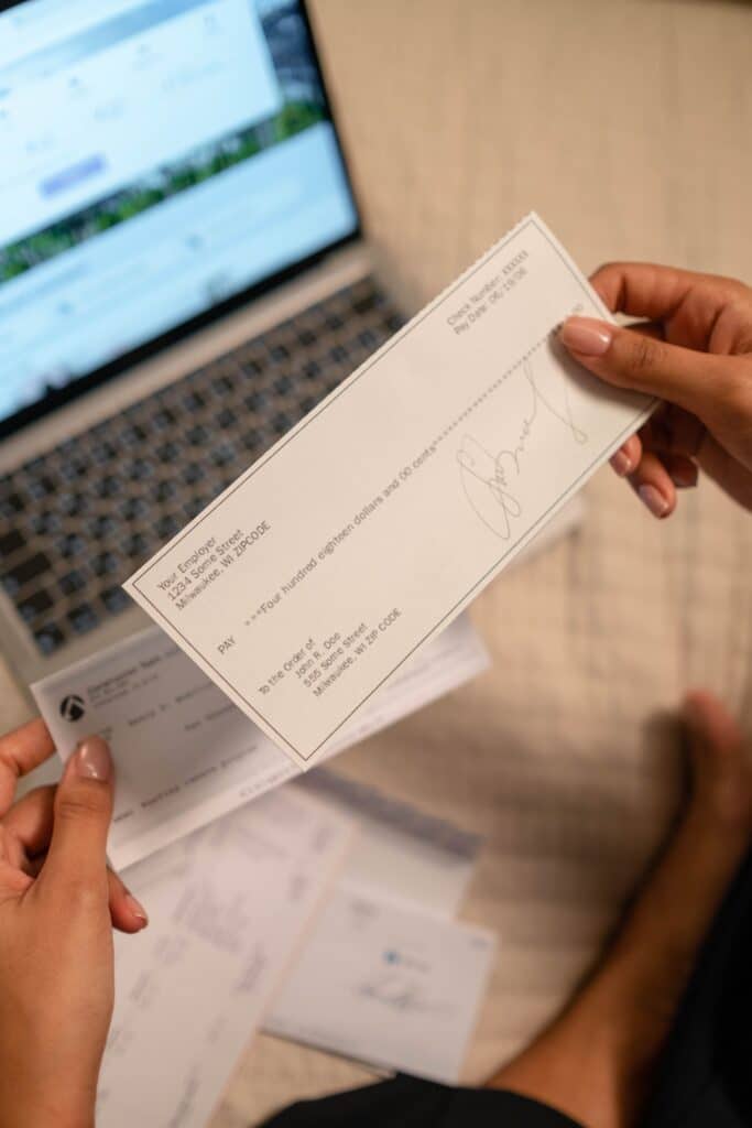 Person holding a signed personal check in front of a laptop with other checks and papers on the desk