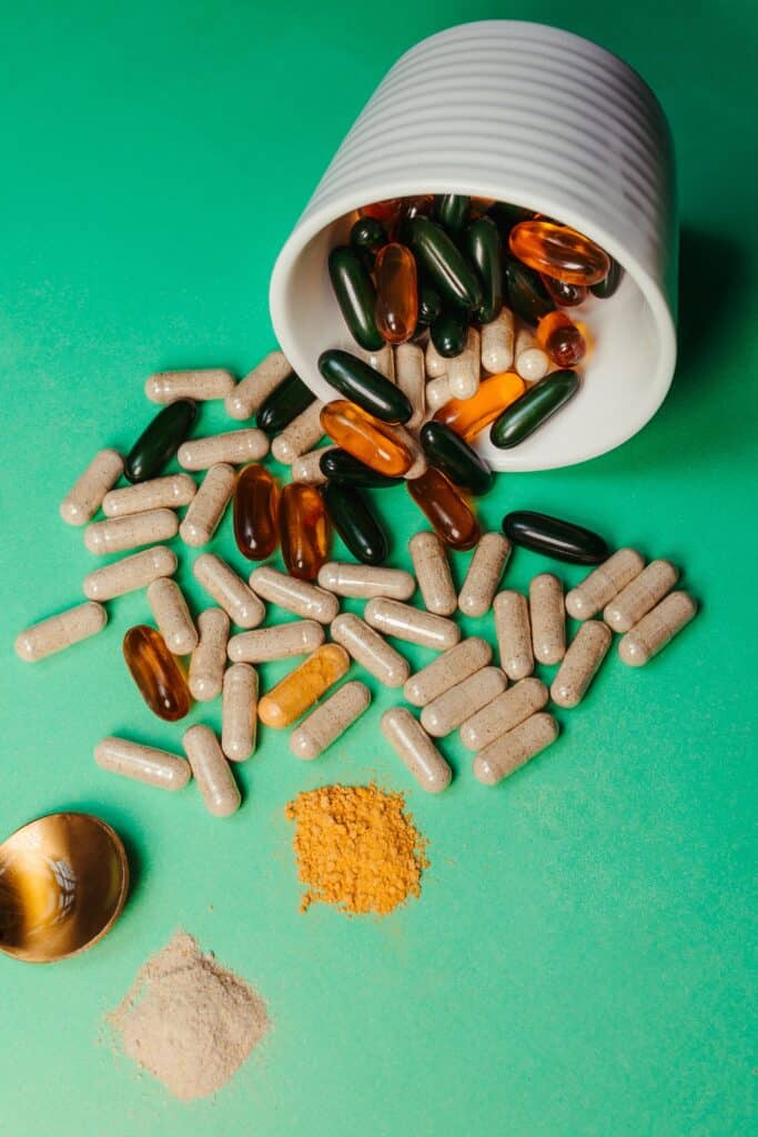 Assorted supplement capsules and softgels spill from a white ribbed cup onto a green surface.