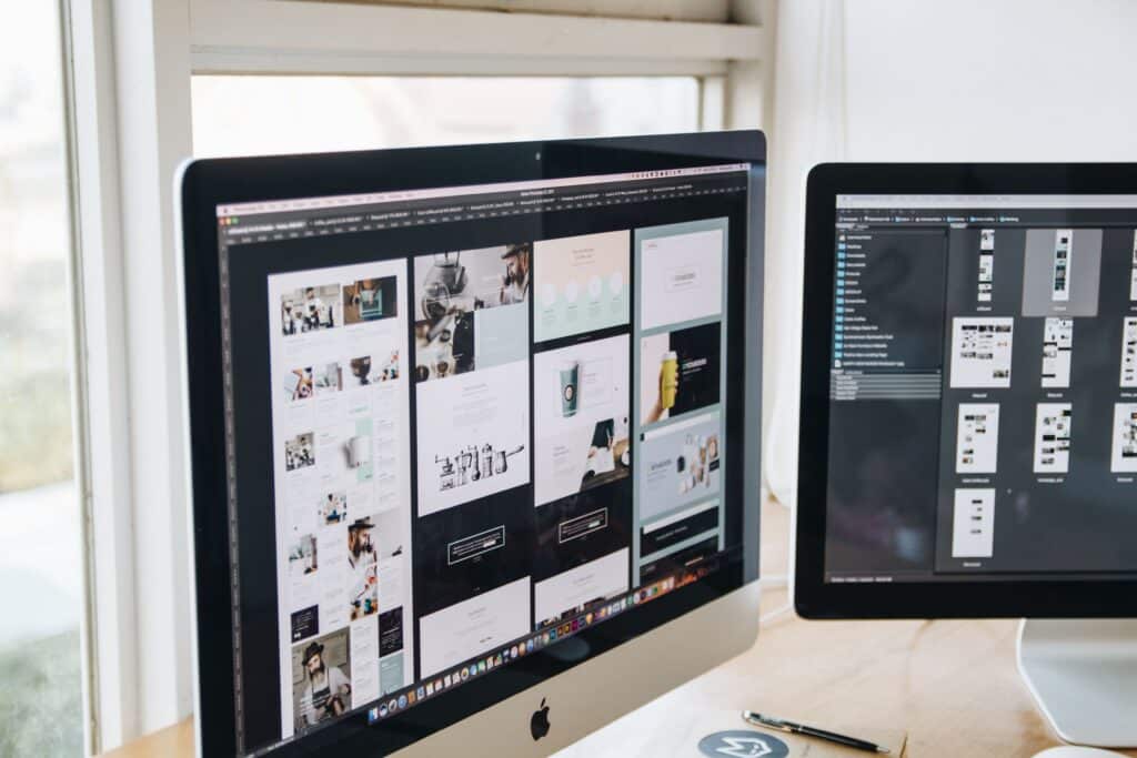 Two monitors on a wooden desk displaying website design templates and layout thumbnails in a design application