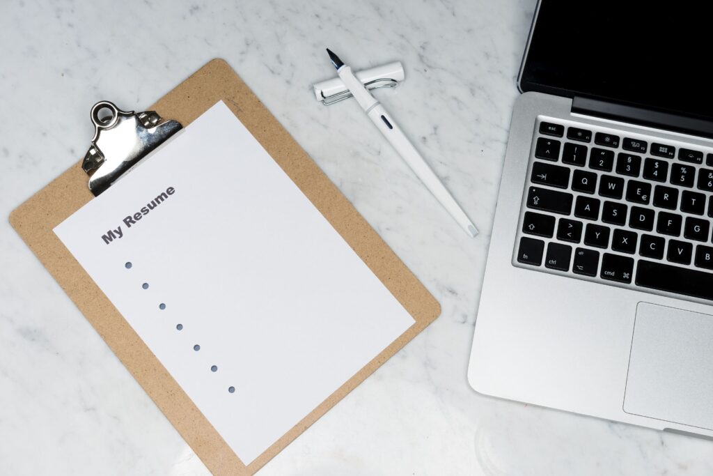 A clipboard holding a blank resume template sits on a marble surface next to an open laptop and two pens.